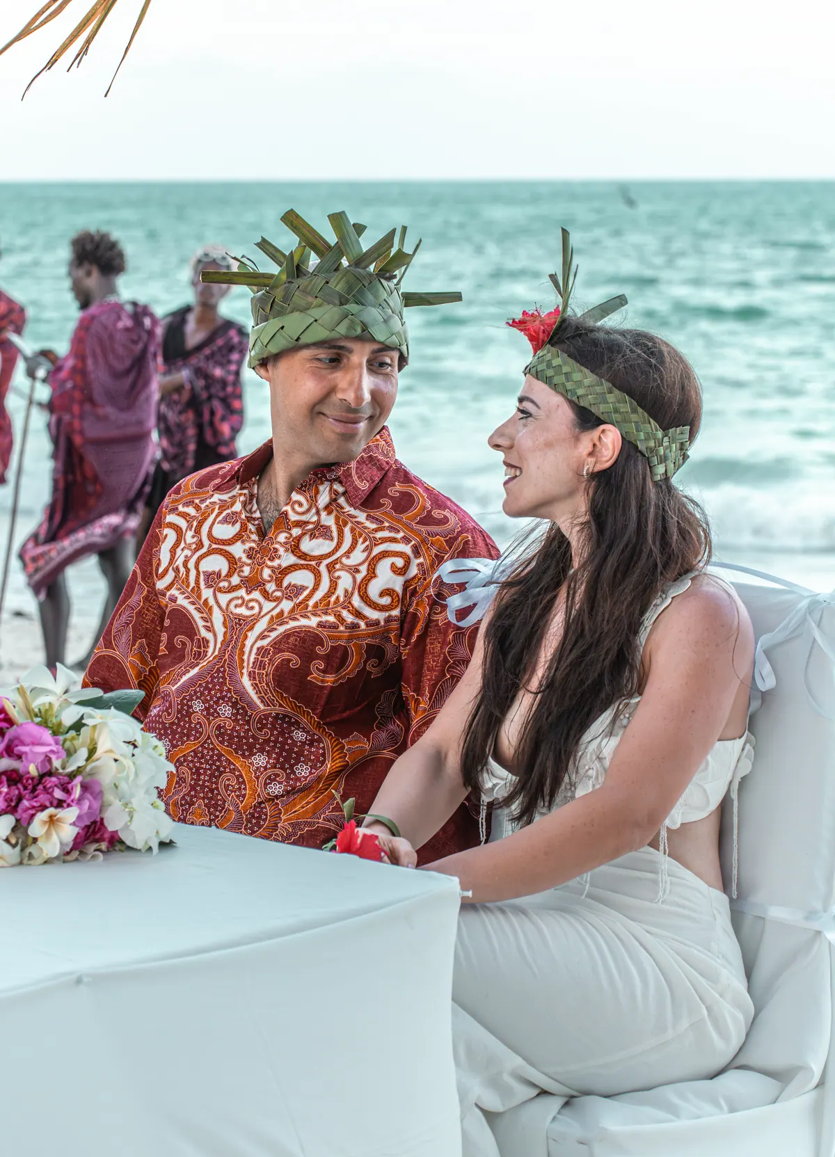 Bride and groom in palm-leaf crowns at the ceremony table by the sea
